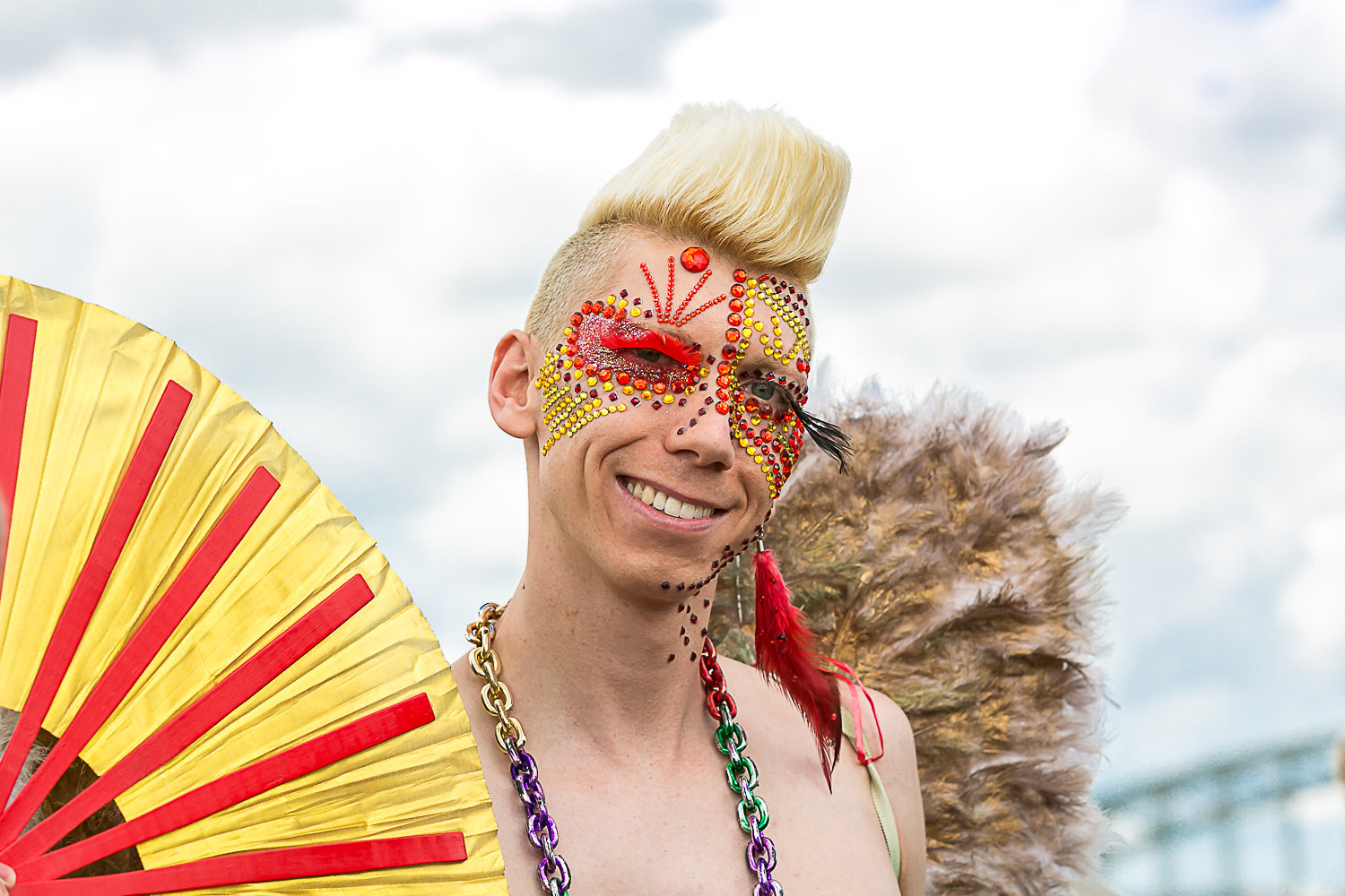 A person poses with a colorful fan and glittering stones on their face in front of the camera.