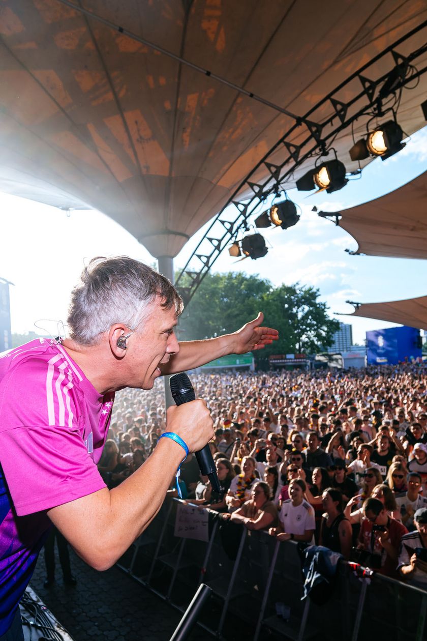 A man stands on a stage in front of a large audience and encourages the people. The sun is shining, clear sky.