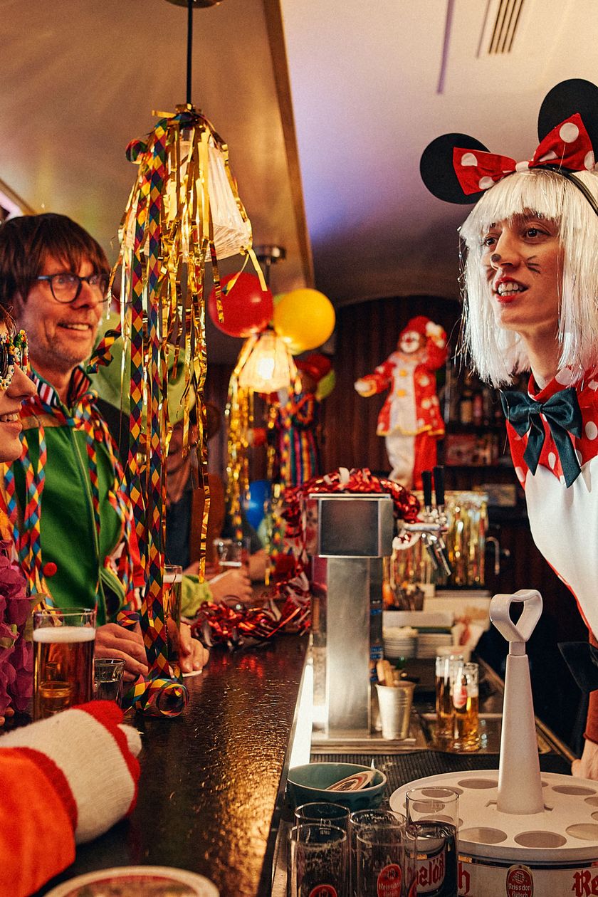 Two disguised women are leaning forward behind a bar counter. They are engaged in conversation with three people standing in front of the bar. Balloons, streamers, and tinsel hang from the ceiling, enveloping the room in a colorful and cheerful atmosphere.
