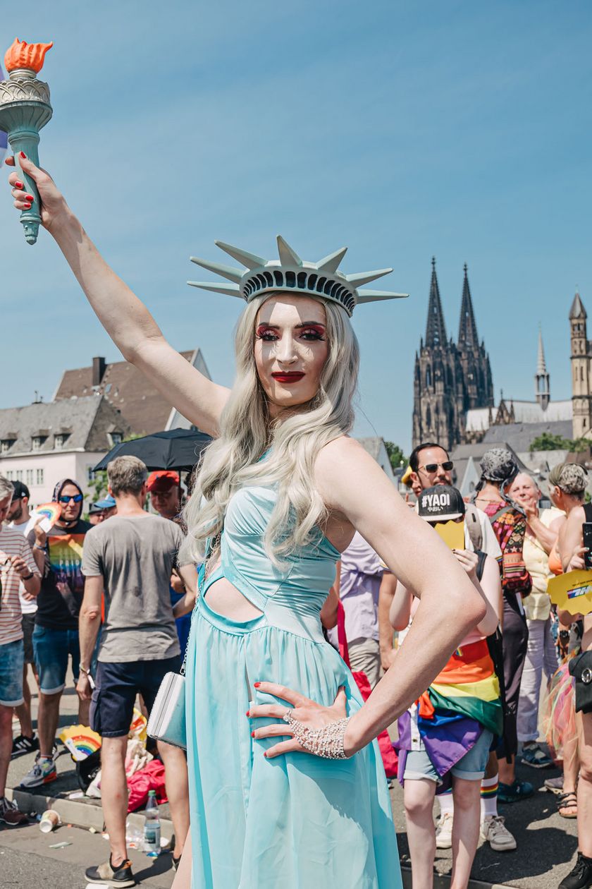 A person dressed as the Statue of Liberty is posing. In the background, people are standing by the roadside.