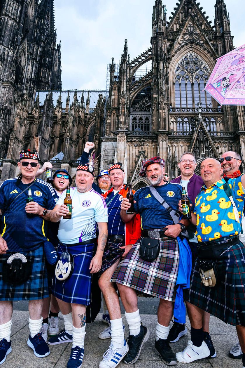 Thirteen men are standing in front of the Cologne Cathedral. Some of them are wearing kilts.
