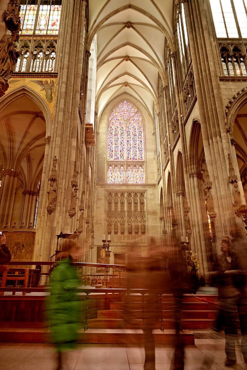 Light breaks through the Richter window in Cologne Cathedral. People move through the interior of Cologne Cathedral.