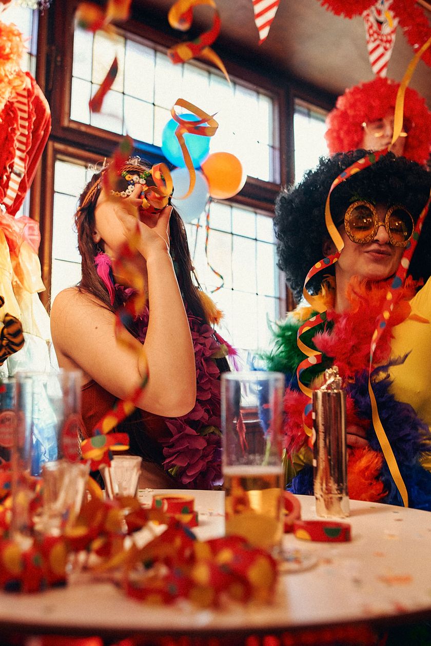 Around a round standing table, four costumed people are gathered. One person is blowing streamers. The room is colorfully and cheerfully decorated.