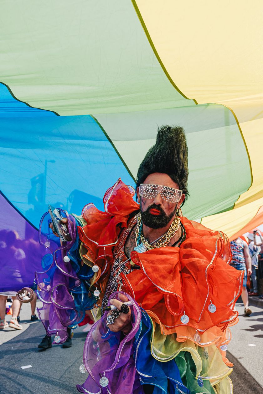 A person in a colorful costume poses. Above them, a rainbow flag is spread out.