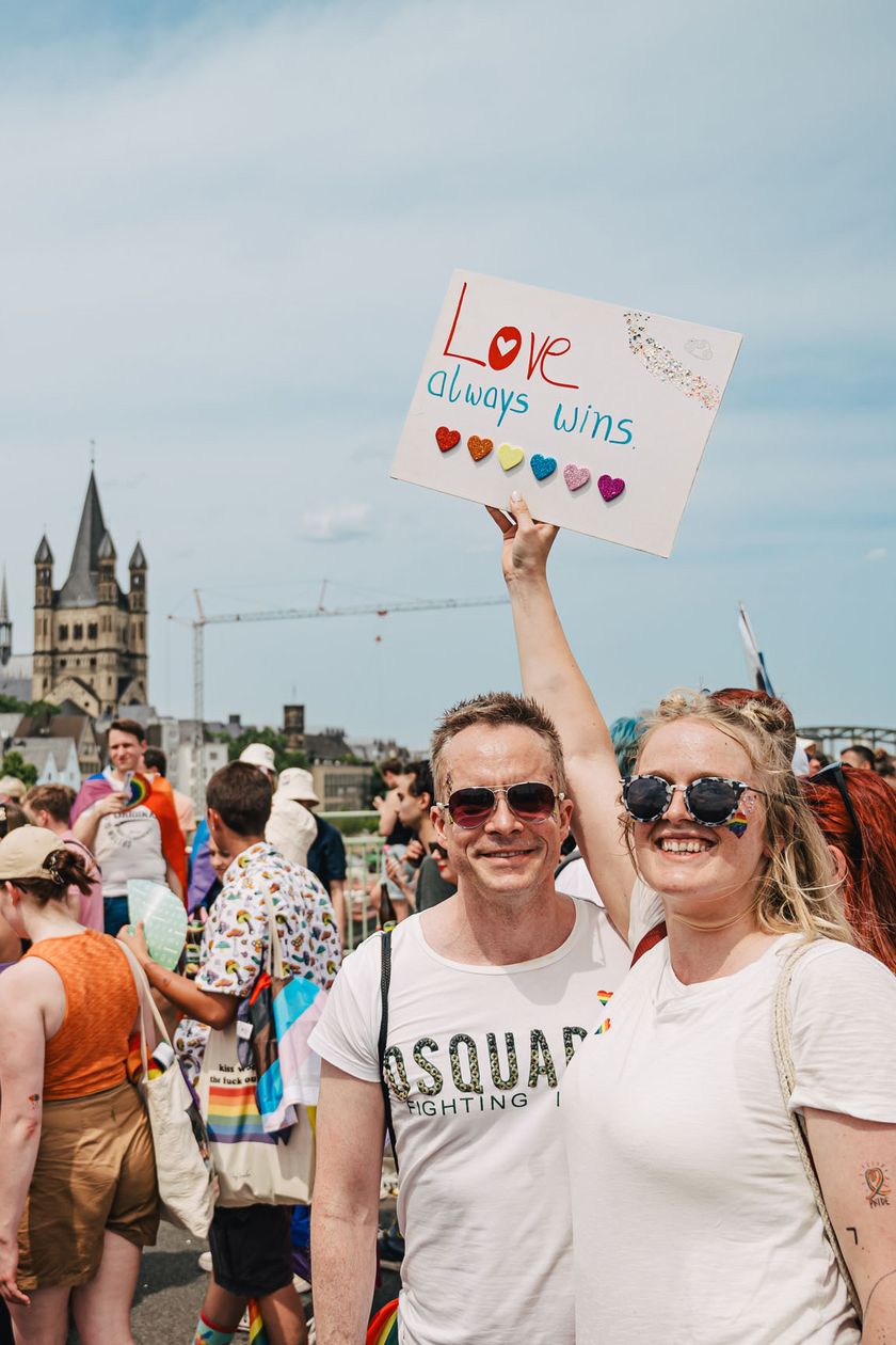 Two people are posing and holding a sign up high. People are walking by, and in the background, the Cologne Cathedral can be seen.