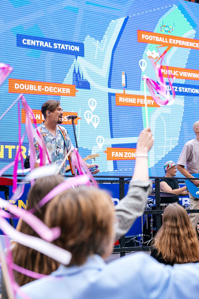 A band is performing in front of an open-air audience. Behind the band is a large LED screen displaying the outlines of Cologne and symbols.