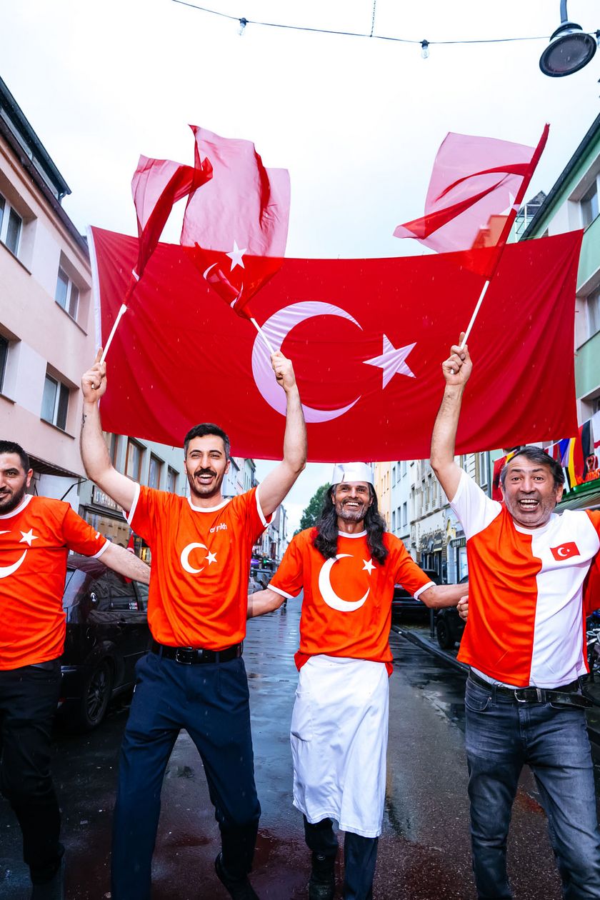 Six men in jerseys are running down a street, holding a large flag in the air.
