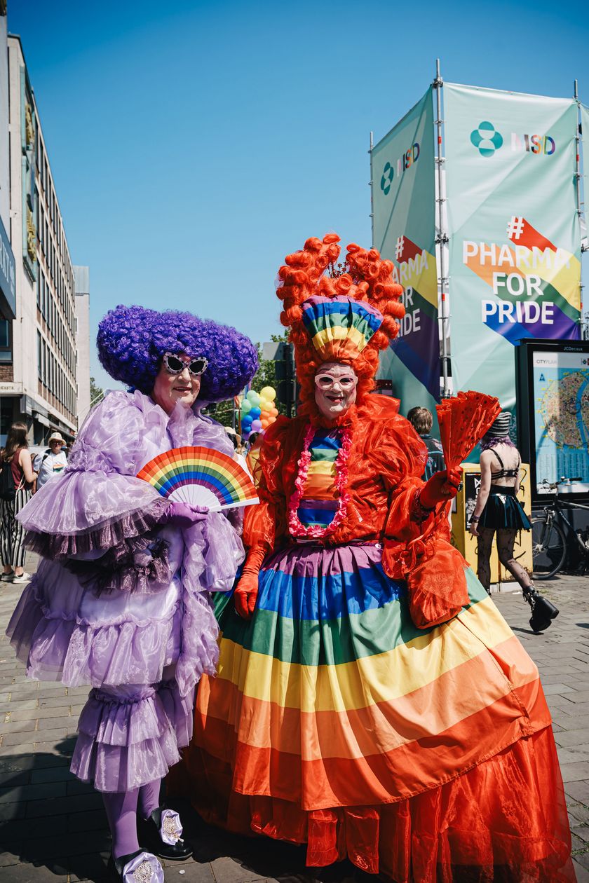 Two people in colorful clothes and extravagant wigs pose under a clear sky.