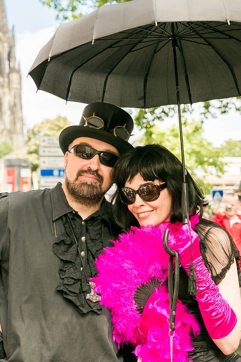 Two people are standing under a black umbrella and wearing extravagant clothing. Cologne Cathedral stands out against the blue sky in the background.
