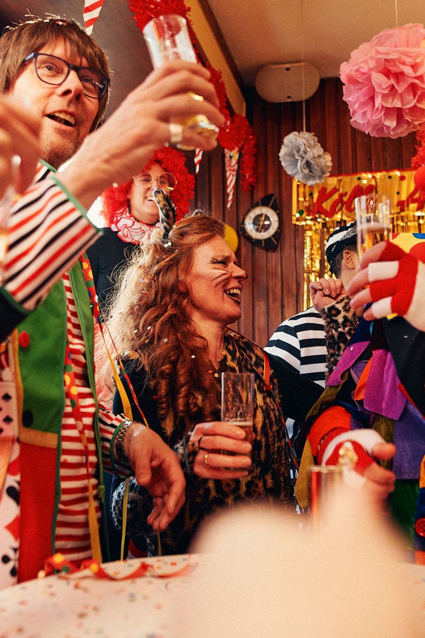A colorful group of people dressed in costumes is holding Kölsch glasses up in the air. The room is decorated with paper garlands and pennants.