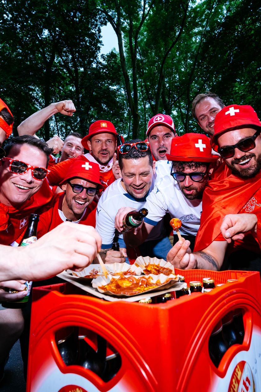 A group of men in red jerseys is standing around a crate of beer. On top of it is a cardboard bowl with currywurst and fries. The men are poking at the dish with a fork and look happy doing it.
