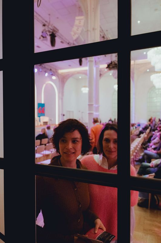 Two people are looking through a glass door. Behind them is a warmly lit hall.
