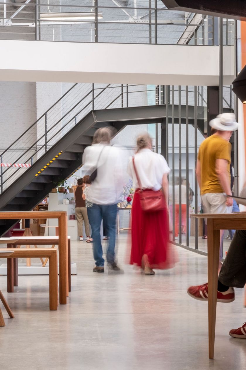 The image shows a brightly lit hall, flooded with light, featuring a bridge connector and a staircase. Designer furniture is placed in the hall, and people are looking around with interest.