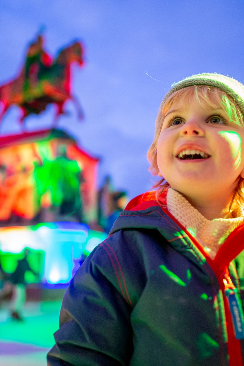 A child with shining, astonished eyes in front of colorful, festive lighting and an equestrian statue.
