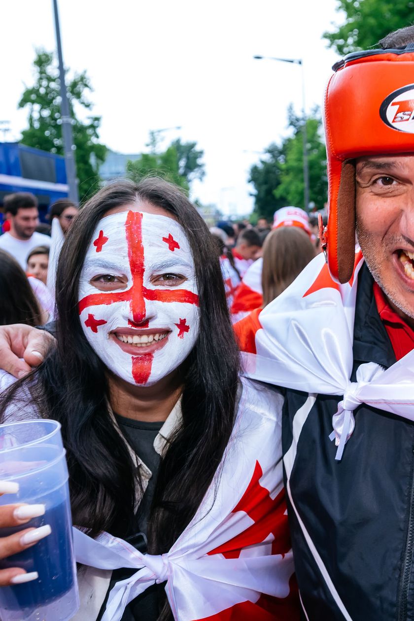 A woman with face paint in the shape of a flag stands smiling next to a man who is holding a boxing glove towards the camera.