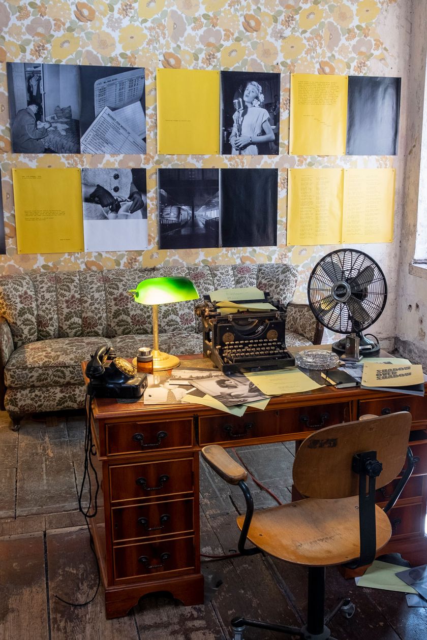 A room with worn wooden floorboards and retro furniture is illuminated with warm light. Black and white photographs are placed on the walls.