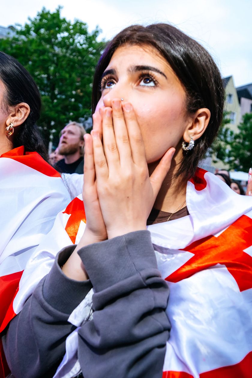 Three women with flags draped over their shoulders look eagerly upwards. One woman has her hands over her face.