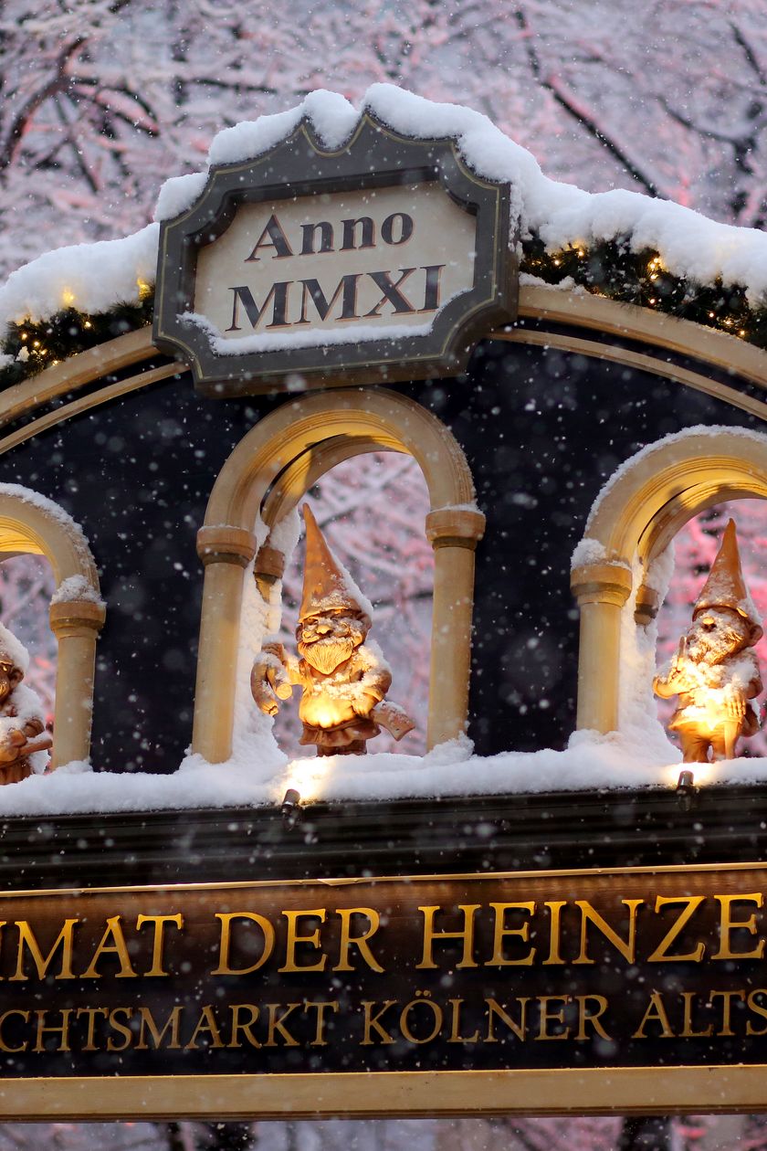 The entrance to the Cologne Christmas market at Heumarkt at dusk. In the image, the decorated archway can be seen with three gnome figures, covered in snow.