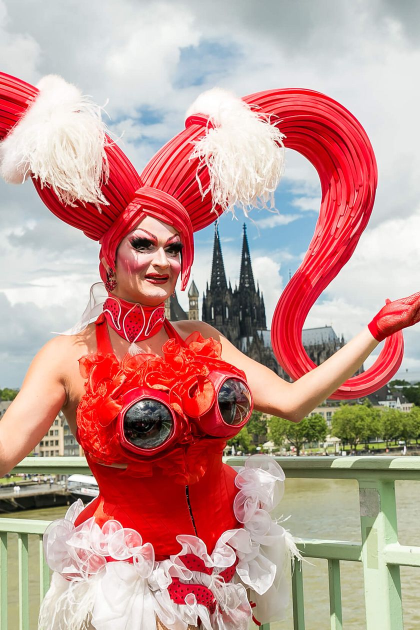 A person in an impressive red costume poses on the Deutz Bridge, with Cologne Cathedral rising in the background.