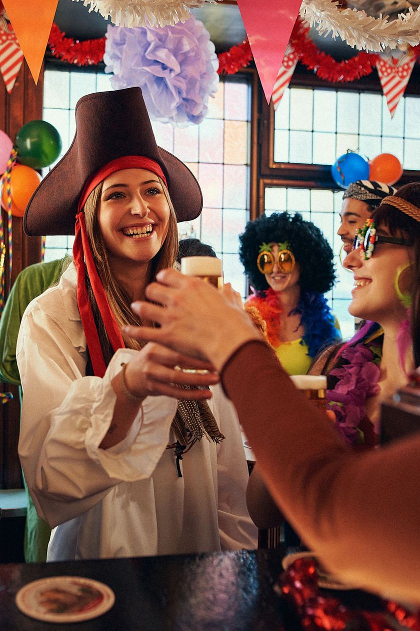 Three costumed individuals are standing at a counter in a colorfully decorated room. A hand is handing a Kölsch glass to the person in the middle.