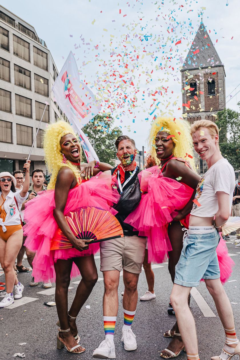 Four people are posing at the Pride parade on the street. Two of them are wearing blonde wigs and pink tulle skirts.