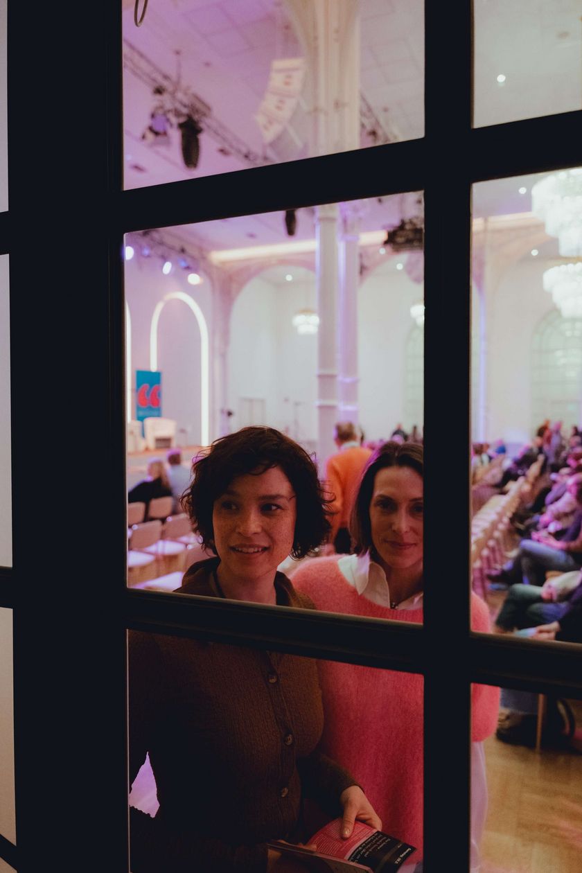 Two people are looking through a glass door. Behind them is a warmly lit hall.