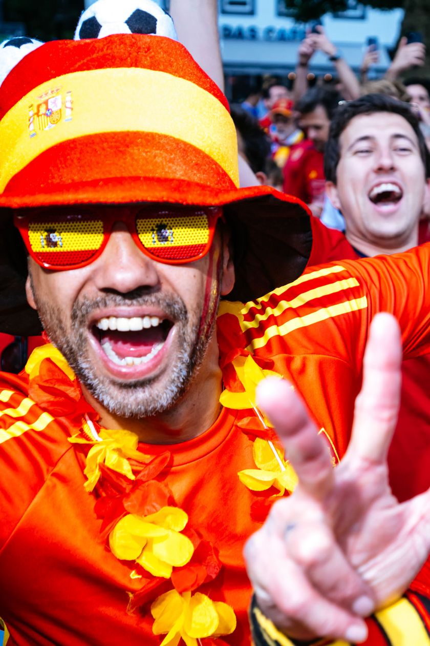 Two men in red jerseys, funny hats, and glasses cheer into the camera and flash the victory sign.