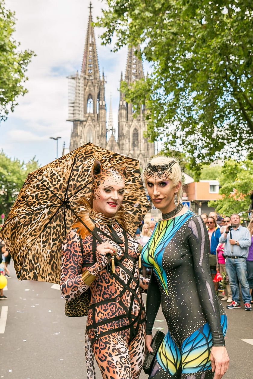 Two people in creative costumes pose on the path of the Pride demonstration. People stand by the roadside and in the background, the Cologne Cathedral stretches.