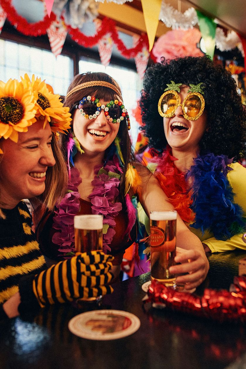 Three women in costumes, with sunflowers in their hair and large round glasses, are standing at a bar counter in a colorfully decorated room, laughing heartily.