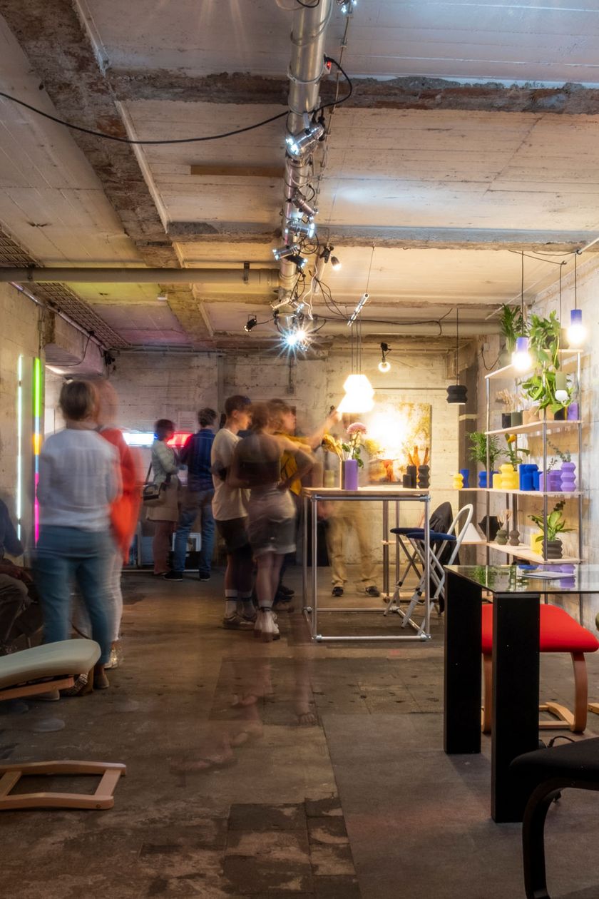 In an underground industrial space with concrete walls, designer furniture pieces and decorative items are scattered around. People are viewing the exhibition.