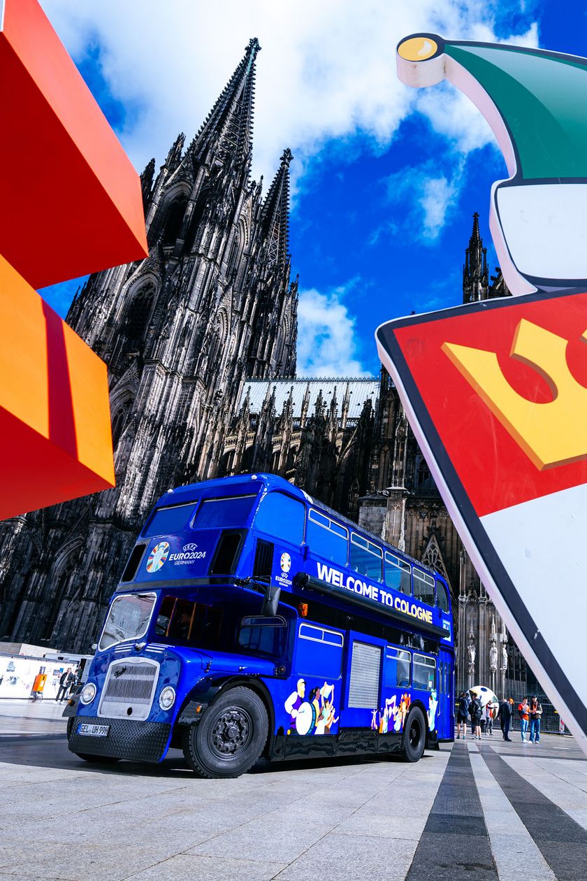 A blue double-decker bus is parked in the square in front of the Cologne Cathedral. Two signs in the foreground flank the image.