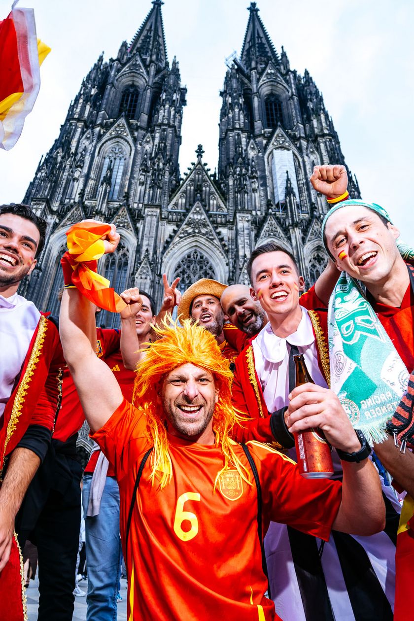 A group of men, some in red jerseys or dressed with wigs, pose jubilantly in front of the Cologne Cathedral.