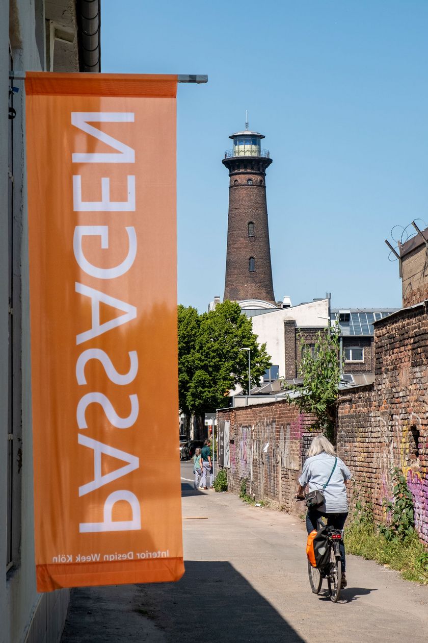 In the image, a side street with a brick wall is visible. A flag with the inscription PASSAGEN stands out prominently. A lighthouse towers in the background.