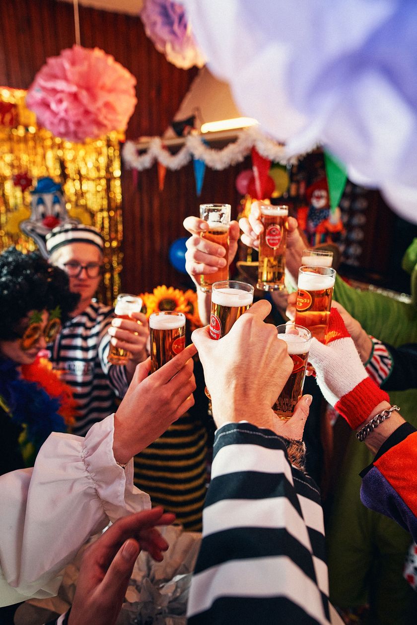 A colorfully decorated room with paper garlands and pennants on the walls and ceiling. In the middle of the image, several hands are clinking Kölsch glasses.