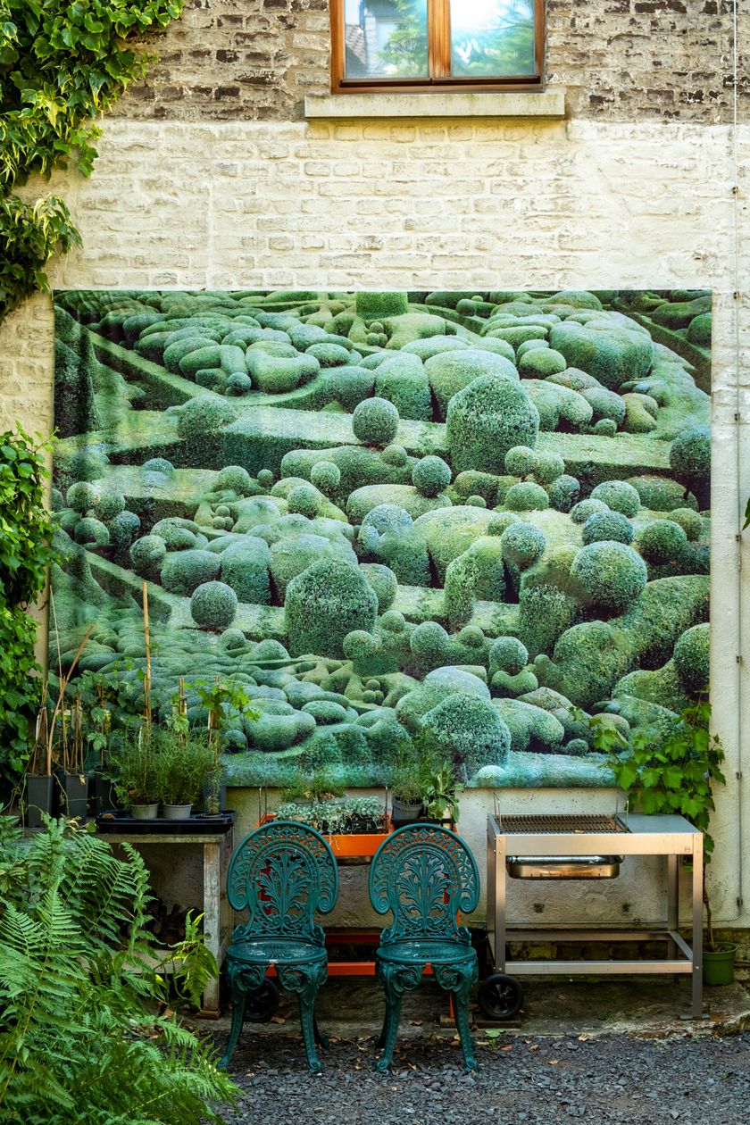 A large painting hangs on the facade of a house in a backyard. In front of it, various pieces of furniture are placed. The color green dominates the photo.